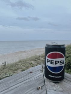 a can of pepsi sitting on top of a wooden table next to the ocean and beach