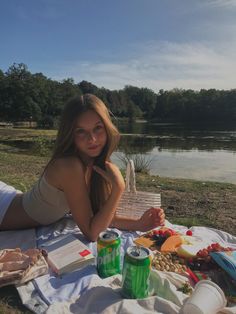 a woman laying on top of a blanket next to a lake with picnic food and drinks