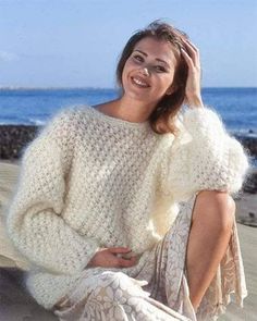 a woman sitting on the beach wearing a white sweater