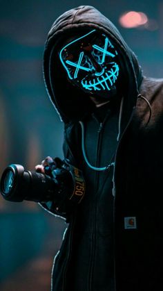 a man in a black hoodie holding a camera and wearing a blue light up mask