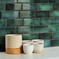 three white cups sitting on top of a counter next to a green tile backsplash