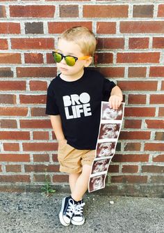 a young boy wearing sunglasses and holding a skateboard in front of a brick wall