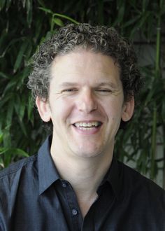 a man with curly hair smiling and wearing a black shirt in front of green plants