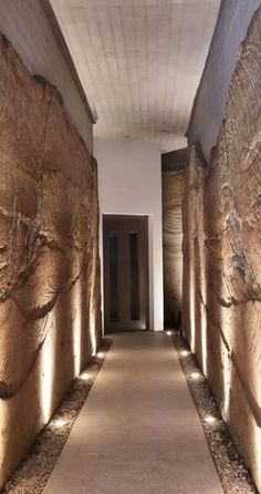 a long narrow hallway with stone walls and lights on either side of the corridor, leading to another room