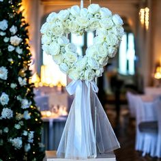 a heart shaped wreath with white roses hanging from it's side in front of a christmas tree