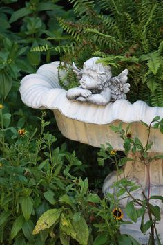 a birdbath in the middle of some plants and flowers, with a little angel figurine sitting on top