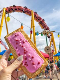 a person holding up a pink frosted pastry in front of an amusement park ride