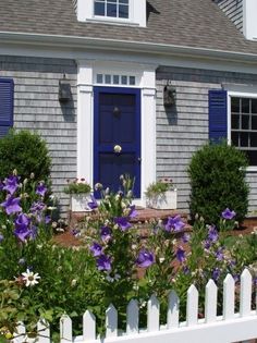 a blue door and white picket fence in front of a gray house with purple flowers