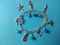 Pretty charm bracelet with a mermaid theme.  Bracelet, which is silver plated, features  a variety of ocean-themed objects, interspersed with crackle glass beads in shades of blue, aqua and green.  The clasp is a cute ship's helm. This is a great gift for girls who love mermaids or a beautiful piece of holiday jewellery for yourself. Turquoise Ocean-inspired Bracelet With Lobster Clasp, Adjustable Ocean-inspired Charm Bracelet With Lobster Clasp, Ocean-inspired Blue Charm Bracelet For Gifts, Adjustable Turquoise Metal Charm Bracelet, Turquoise Metal Charm Bracelet Gift, Turquoise Metal Charm Bracelet As Gift, Silver Beaded Ocean-inspired Bracelets, Ocean-inspired Silver Beaded Bracelets, Blue Metal Charm Bracelet With Lobster Clasp