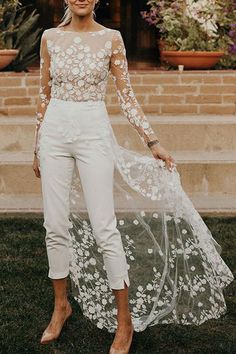 a woman standing in the grass wearing white pants and a sheer top with flowers on it