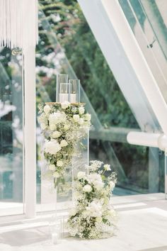 two tall vases with white flowers and greenery on the floor next to each other