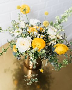 a vase filled with yellow and white flowers on top of a gold table next to a brick wall