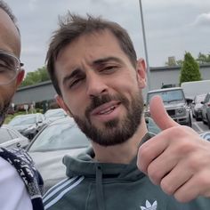 two men standing next to each other in front of parked cars giving the thumbs up sign