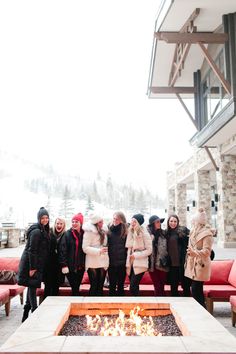 a group of people standing around a fire pit in front of a building with snow covered mountains behind them