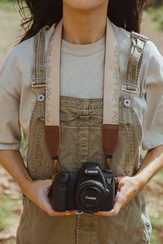 a woman in overalls holding a camera up to her chest and smiling at the camera