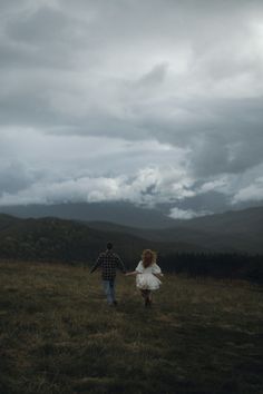 a man and woman holding hands while walking through a field with mountains in the background