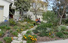 two people sitting on a bench in front of a house with flowers and plants around it