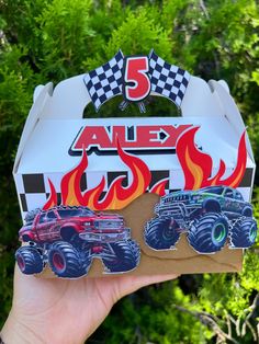 a hand holding up a box that has monster trucks on it and flames in the background