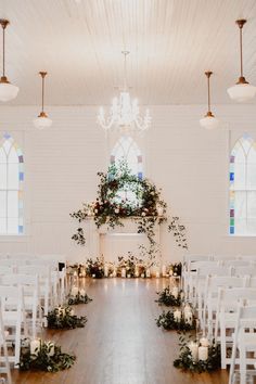 an indoor wedding venue with white chairs and greenery on the aisle, candles in front of them