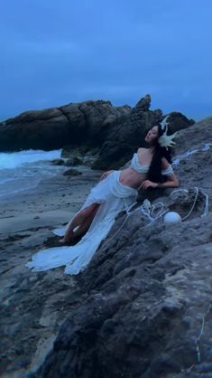 a woman laying on top of a rocky beach next to the ocean with her hair blowing in the wind