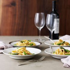 three white bowls filled with pasta and asparagus next to two glasses of wine