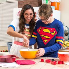 two people in the kitchen preparing food for their family's dinner party, one is wearing a superman shirt and the other is holding a yellow bowl