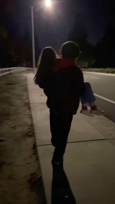 two people walking down a sidewalk at night with the street light shining on them and one person holding onto his back