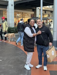 two people standing next to each other in front of a building with a dog on the sidewalk