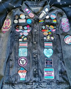 A dark grey denim jacket adorned with patches and pin badges with transgender, bisexual, nonbinary and gay themes down the front of the jacket, on the pockets, on the lapel and on the shoulders. Jean Jacket With Pins Outfits, Pride Battle Jacket, Battle Jackets Punk, Denim Jacket With Patches And Pins, Emo Denim Jacket, Jean Patch Jacket, Battle Vest Patches, How To Make A Battle Jacket