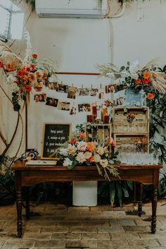 a wooden table topped with lots of flowers and plants next to a wall covered in pictures