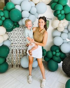 a woman holding a small child in front of a backdrop with green and white balloons