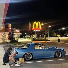 a man sitting on the ground next to a blue car in front of a mcdonald's