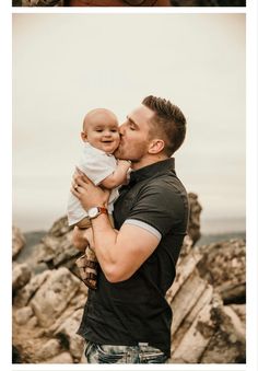 a man holding a baby in his arms while standing on some rocks near the ocean