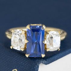 A JbyG trilogy ring with the most delicious sapphire we've come across in a while! That delicious royal blue velvety color in the center is irresistible, flanked by chunky, rustic elongated old mine cut diamonds. Together, they give you tons of color, sparkle, and spread across the top of one's finger. All set in yellow gold, this is one of our favorite original rings to come out of production as of late! 18kt yellow gold Size 6 and fully resizable Sapphire measures 9.87 x 5.53 x 3.75mm, heated Modern Mens Rings, Diamond Trilogy Ring, Art Jewelry Earrings, Trilogy Ring, Antique Watches, Antique Engagement, Mens Band, Vintage Band, Men's Rings