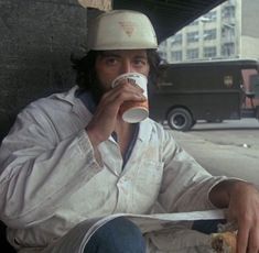 a man wearing a hard hat and drinking from a paper cup while sitting on the ground