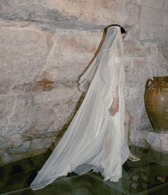 a woman in a white dress and veil standing next to a vase