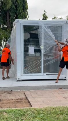 two men in orange vests moving a white building