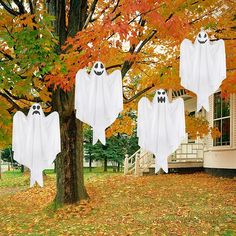 three ghost decorations hanging from a tree in front of a house with fall leaves on the ground