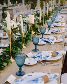 the table is set with blue and white plates, silverware, candles and greenery