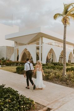 a bride and groom walking down the sidewalk in front of a white building with palm trees