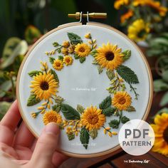 someone is holding up a embroidery project with sunflowers in the center and green leaves around it