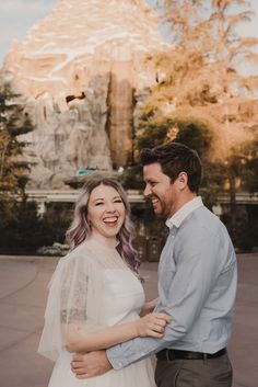 a man and woman standing next to each other in front of a building with a mountain behind them