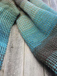 a blue and brown knitted scarf laying on top of a wooden floor next to a piece of wood