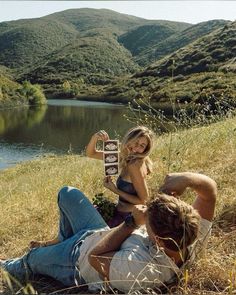 a man holding a skateboard next to a woman laying on the ground in front of a lake