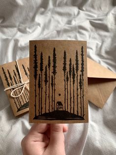 a hand holding up a card with trees on it and a house in the woods