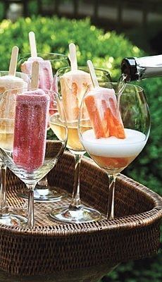 wine glasses filled with different types of drinks sitting on top of a wooden tray in front of bushes