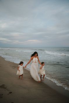 a mother and her two daughters walking on the beach holding hands with each other as they walk towards the water