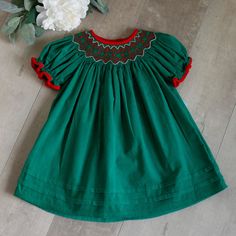 This Emerald Green Baby Girl's Smocked Dress Is Precious!! The Dress In Done In 100% Cotton Pin Wale Corduroy. The Lovely Emerald Green Color Has A Red Matching Corduroy Trim Piping At The Sleeve Hem And The Neckline. The Smocking Is Adorned With Red, Green And White Embroidery. Sweet Red Roses Adorn Adorn The Front Of The Smocking. The Hem Of The Dress Has 2 French Tucks Adding Another Element Of Style To This Classic Bishop/Float Style Dress. Upper Button Back Closure. Perfect For Southern War Red Hand Smocked Dress, Smocked Christmas Outfits, Baby Christmas Dress Green, Smocked Christmas Dresses, Girls Smocked Christmas Dress, Smocked Baby Girl Dresses, Smocked Bishop Dress, Swan Dress, Girls Smocked Dresses