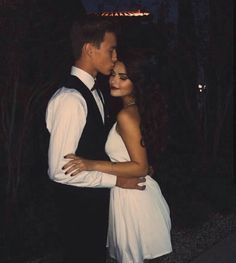 a man and woman standing next to each other in front of some trees at night