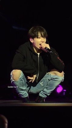 a young man sitting on top of a stage holding a microphone in his hand and wearing ripped jeans